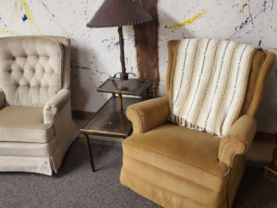 one yellow and one beige easy chair with a sidetable in between against a paint-speckled white wall